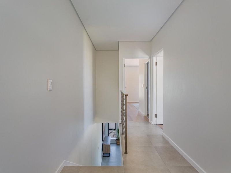 A bright, neutral-toned hallway featuring tiled flooring and a modern metal balustrade overlooking the staircase, leading to adjacent rooms.