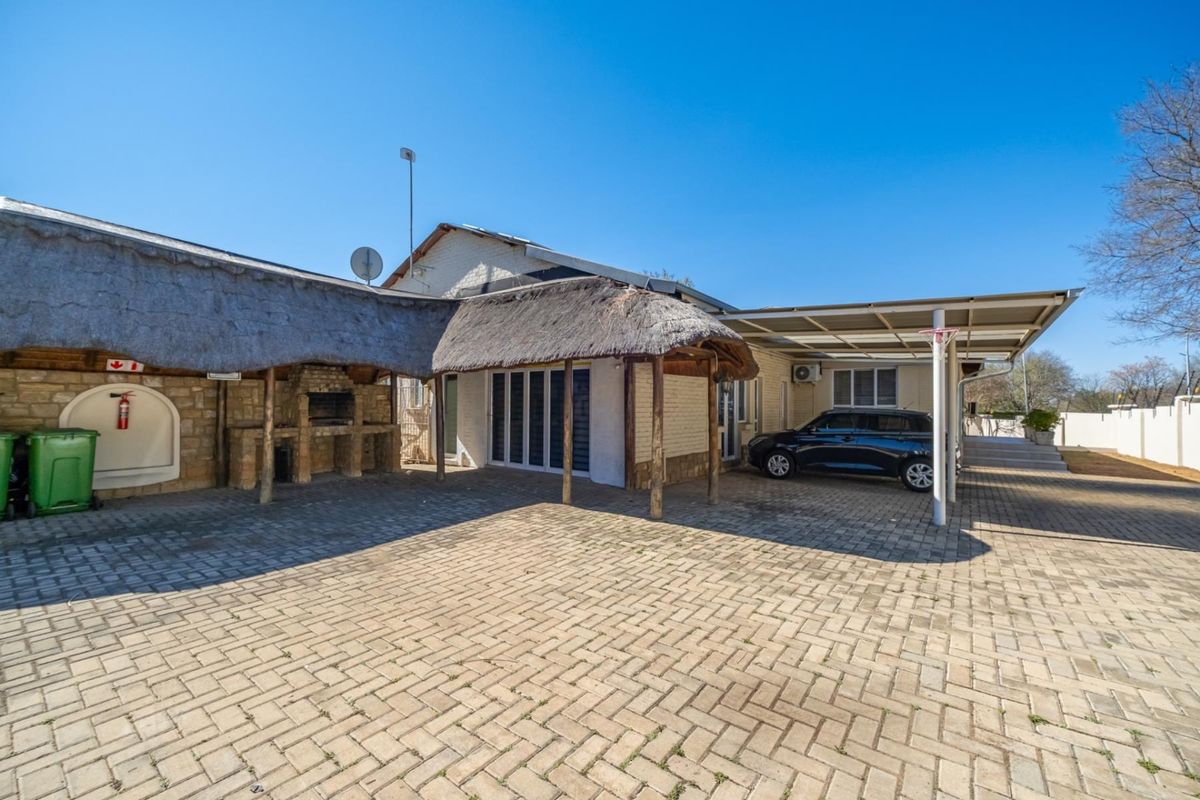 Rear exterior view featuring a paved courtyard, a thatched-roof entertainment area with a built-in braai, and a covered carport.