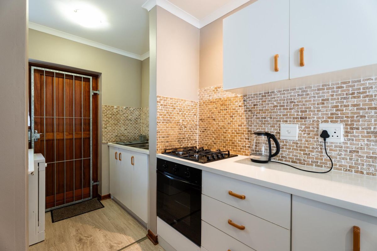 A compact kitchen featuring white cabinetry, a gas hob, built-in oven, and a decorative mosaic tile backsplash, with direct access to a security gate.