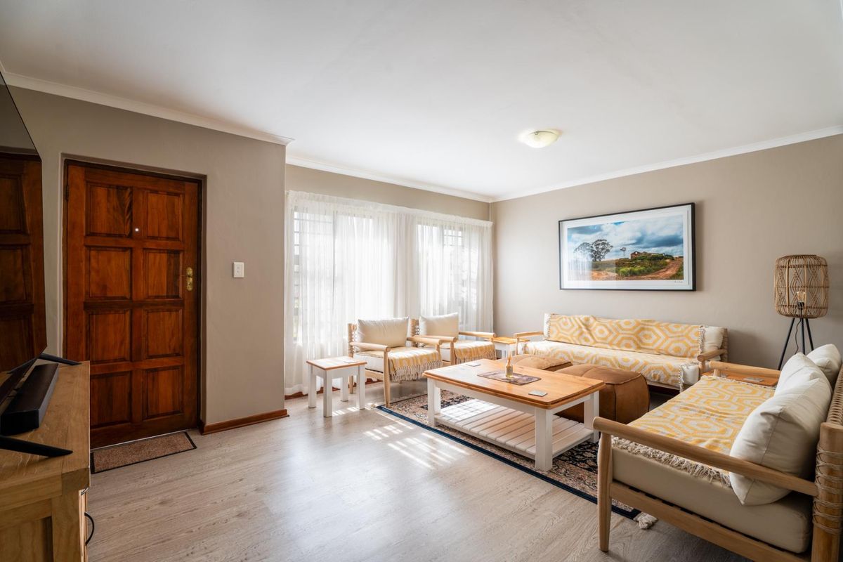 A well-maintained living room featuring light-toned laminate flooring, neutral wall paint, and a solid timber entrance door with natural light from large windows.