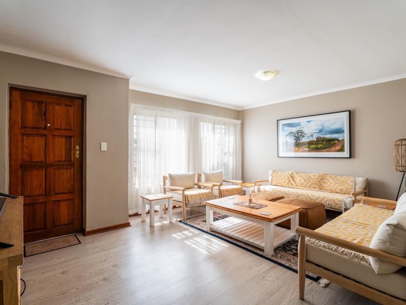 A well-maintained living room featuring light-toned laminate flooring, neutral wall paint, and a solid timber entrance door with natural light from large windows.