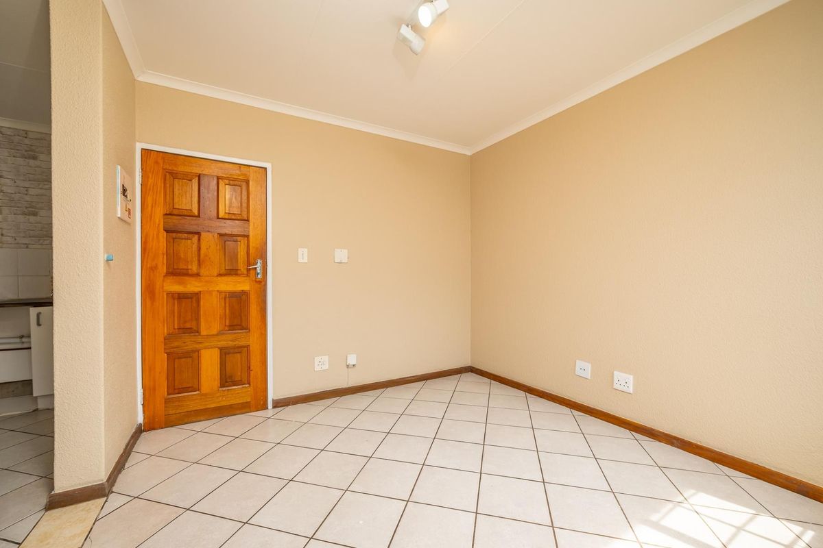 An empty living area featuring neutral wall paint, tiled flooring, and a solid wooden door, with a glimpse of the adjacent kitchen area.