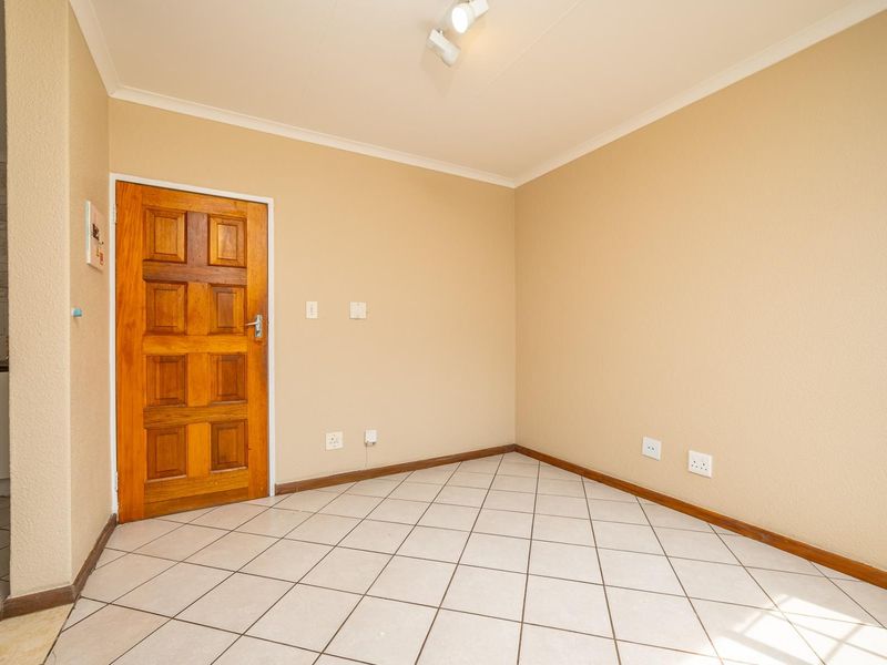An empty living area featuring neutral wall paint, tiled flooring, and a solid wooden door, with a glimpse of the adjacent kitchen area.