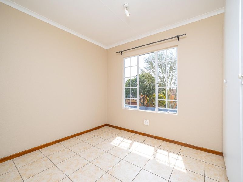 A bright, empty bedroom featuring neutral tiled flooring, a built-in cupboard, and a window with security bars, offering a clean and versatile living space.