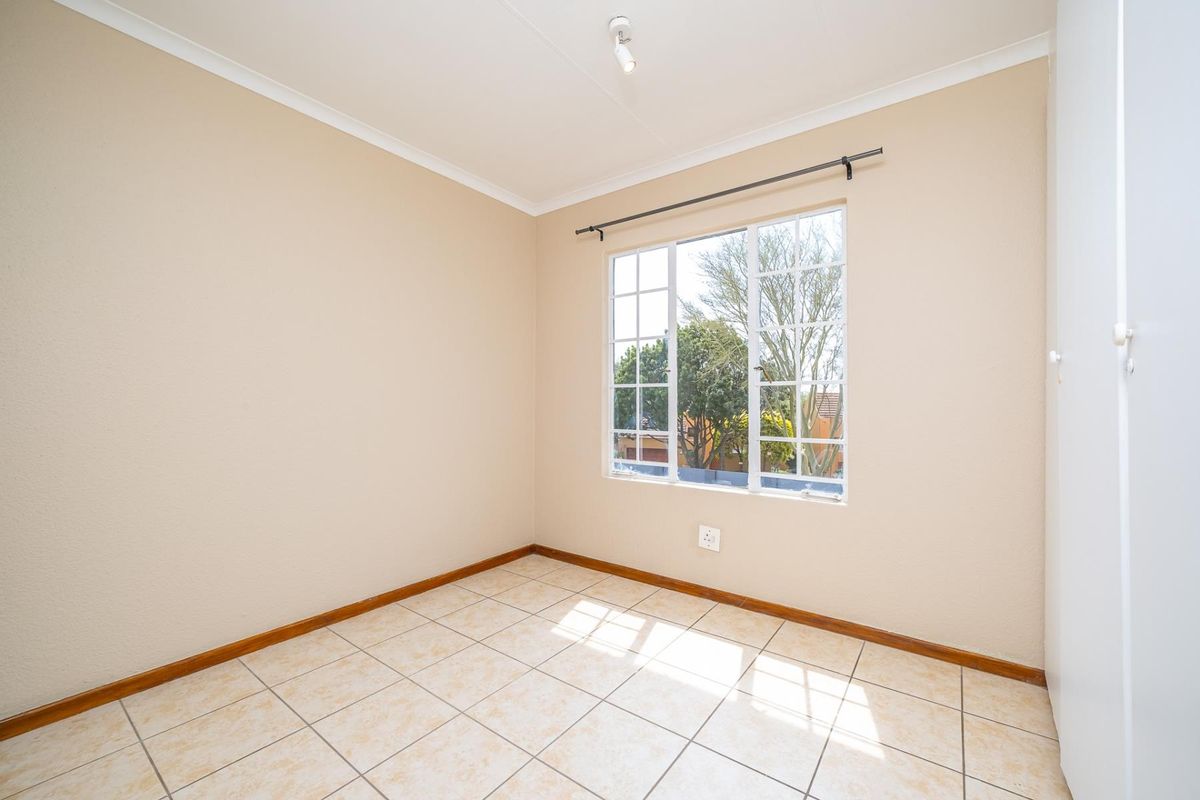 A bright, empty bedroom featuring neutral tiled flooring, a built-in cupboard, and a window with security bars, offering a clean and versatile living space.