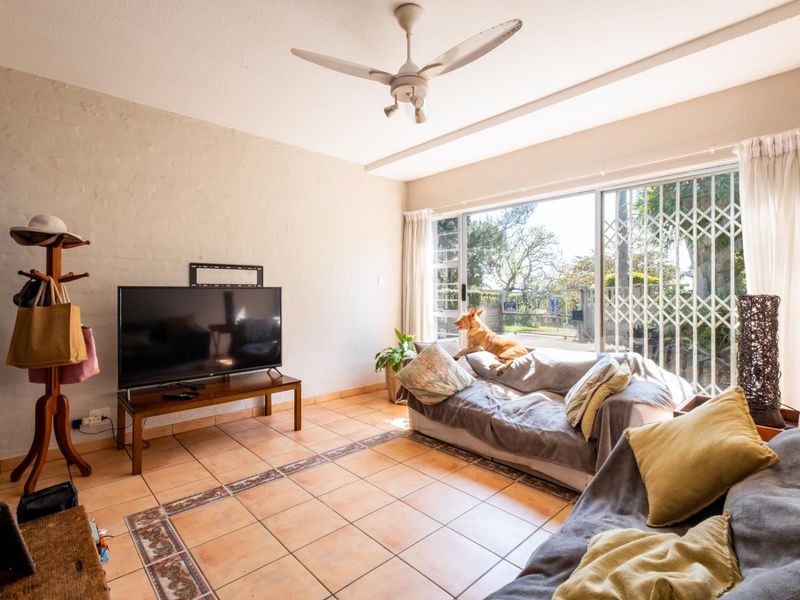 A sunlit living room featuring ceramic tiled flooring, a ceiling fan, and large sliding glass doors with security gates leading to the exterior.