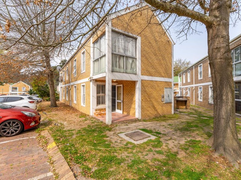 Rear exterior view of a two-story brick apartment building featuring a small patio area with a built-in braai and established trees.