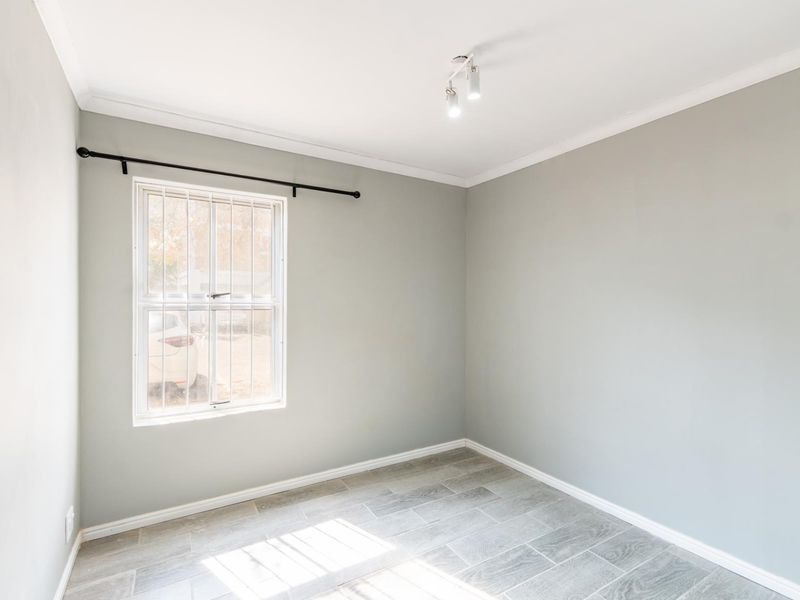 A bright, empty bedroom featuring neutral grey walls, tiled flooring, and a window fitted with security bars.