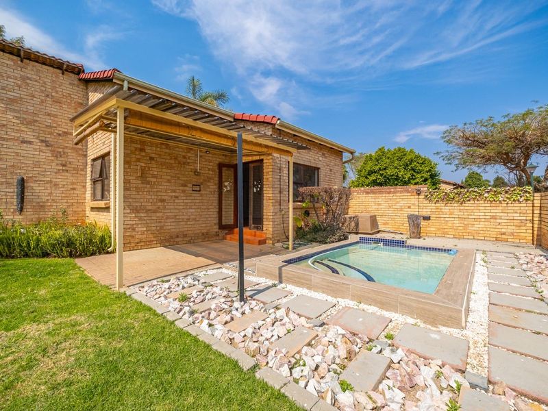Rear exterior view featuring a brick house, covered patio area, and a small swimming pool set within a landscaped garden with paved pathways.