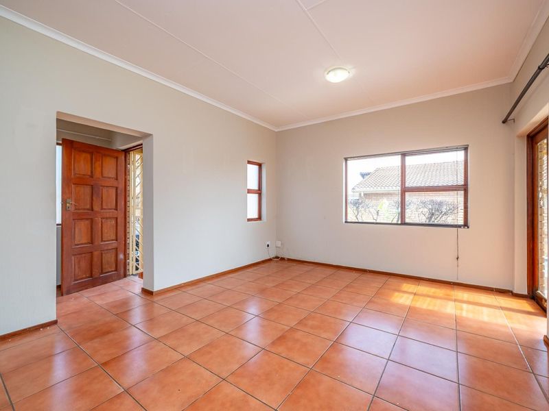 A bright, empty living room featuring terracotta-style floor tiles, neutral walls, and natural light through aluminium-framed windows.