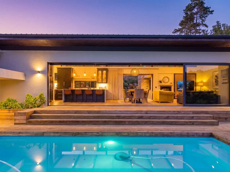 A beautifully lit rear exterior view featuring a swimming pool, expansive timber decking, and large sliding doors opening into an open-plan living and kitchen area.