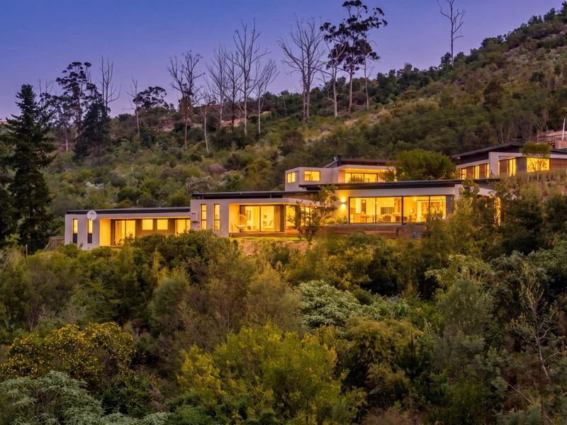 A stunning multi-level modern residence nestled into a hillside, featuring expansive glass facades, timber decking, and integrated outdoor lighting at dusk.