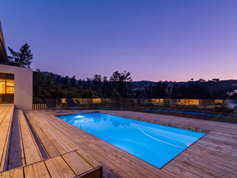A beautifully illuminated swimming pool area featuring extensive timber decking and a modern glass balustrade, set against a scenic evening landscape.