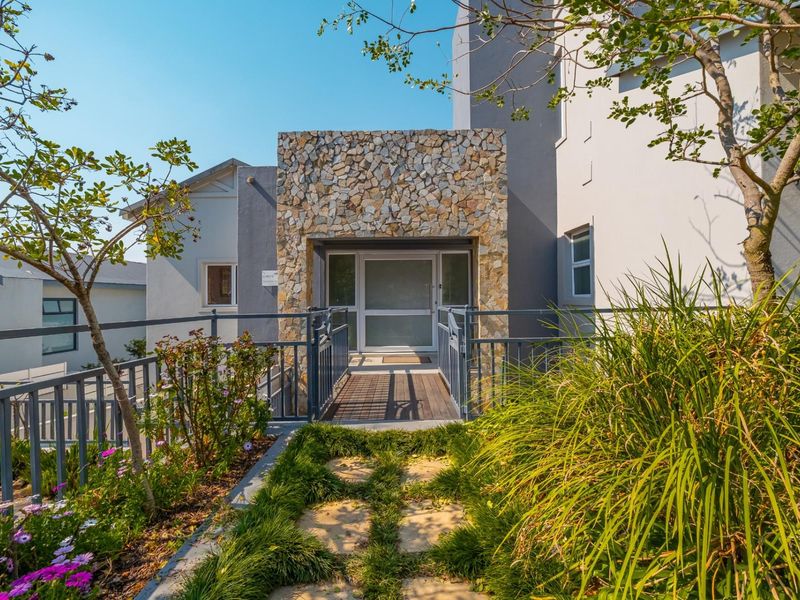 Modern front entrance featuring a stone-clad facade, timber decking walkway, and metal railings, leading to a glass-panelled front door.