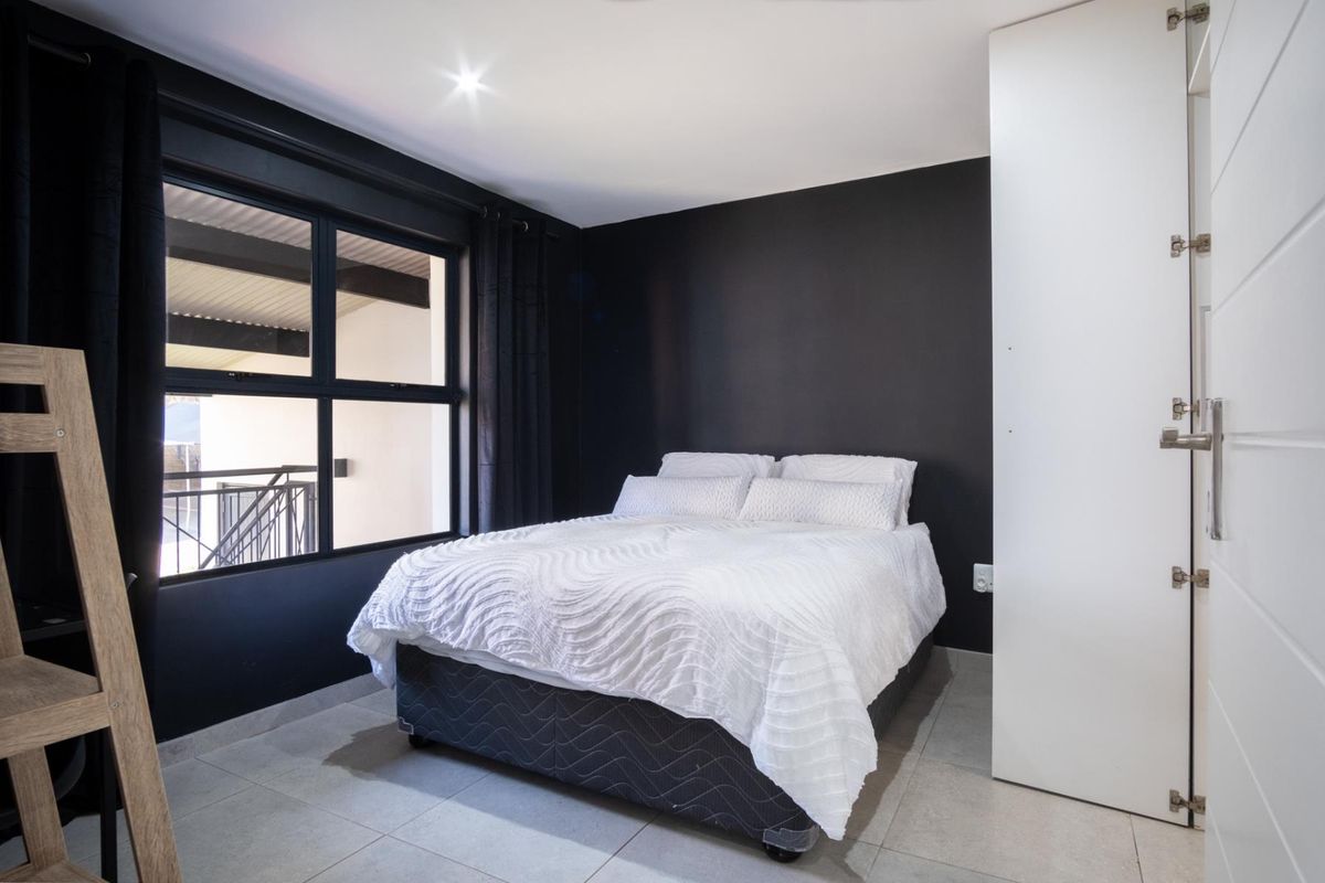 A modern bedroom featuring dark accent walls, tiled flooring, a built-in cupboard, and large aluminium-framed windows allowing natural light.