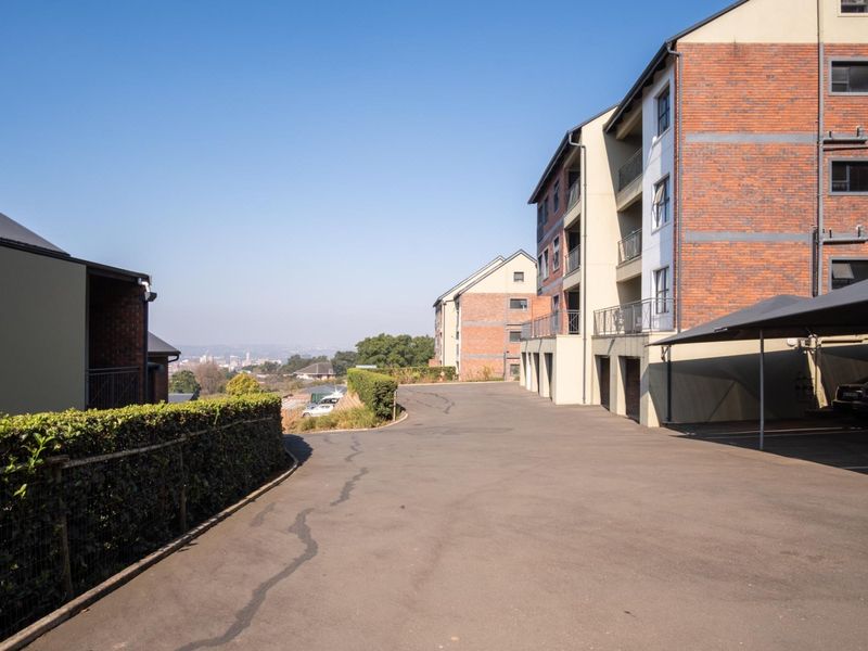 Exterior view of the apartment complex featuring brick facade buildings, covered parking bays, and an asphalt driveway with surrounding landscaped hedges.