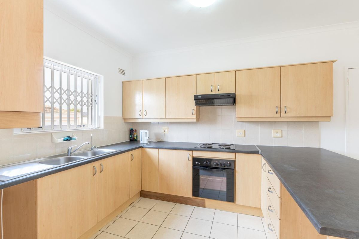A functional kitchen featuring light wood-finish cabinetry, dark laminate countertops, built-in oven, electric hob, and tiled flooring with a window security gate.