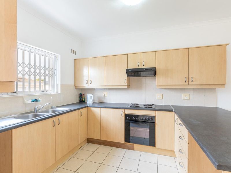 A functional kitchen featuring light wood-finish cabinetry, dark laminate countertops, built-in oven, electric hob, and tiled flooring with a window security gate.