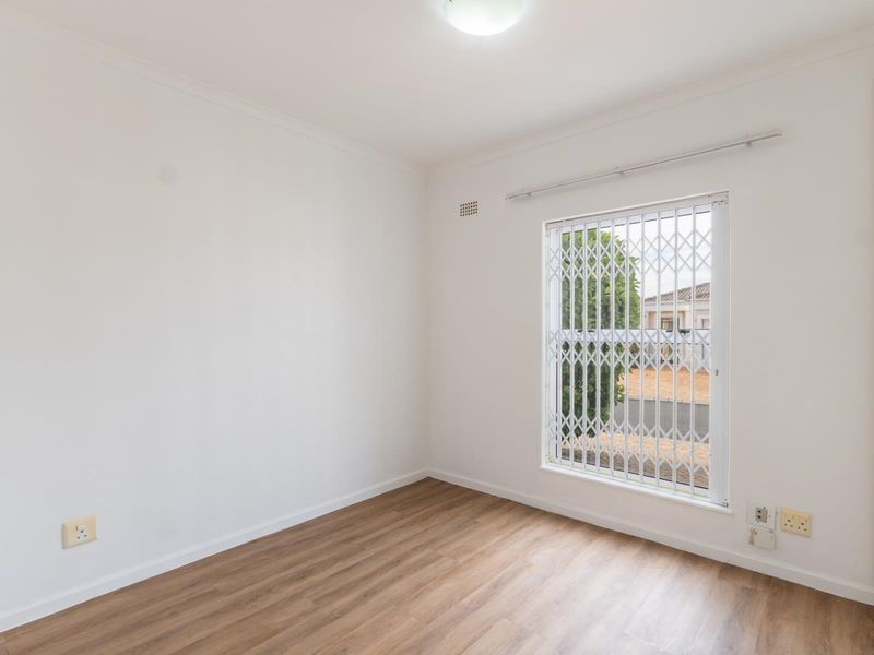 A bright, empty bedroom featuring modern laminate flooring, a built-in cupboard, and a window fitted with a security gate.