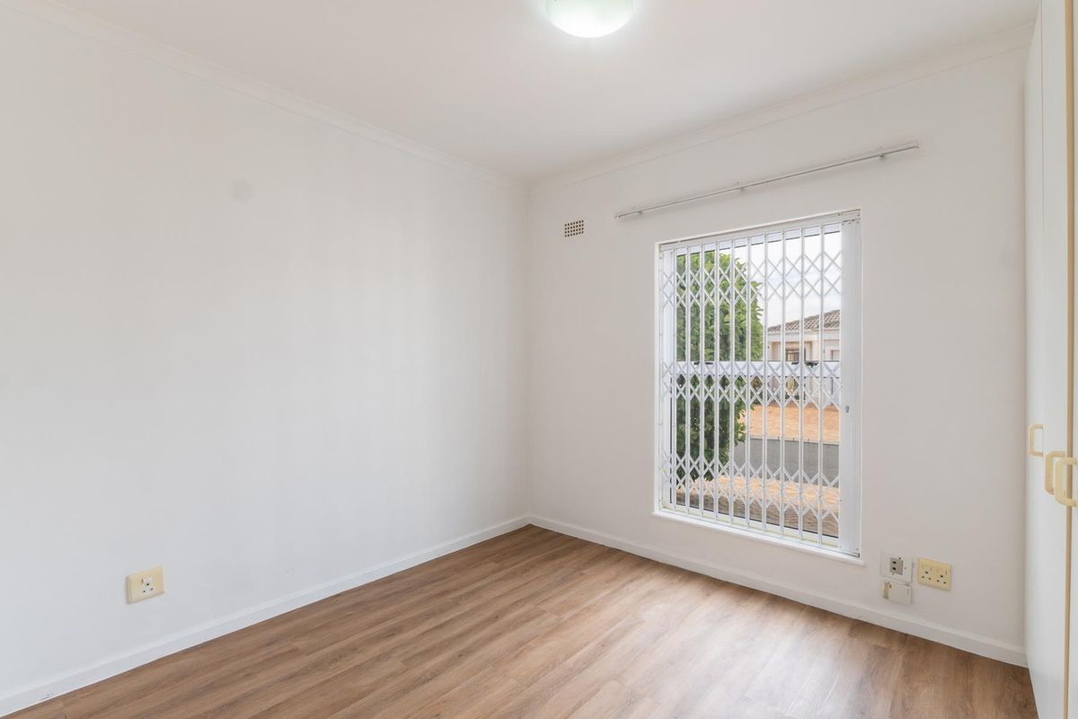 A bright, empty bedroom featuring modern laminate flooring, a built-in cupboard, and a window fitted with a security gate.