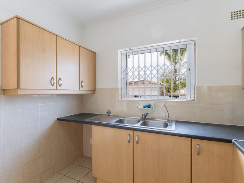 A functional scullery area featuring light wood cabinetry, dark laminate countertops, a double stainless steel sink, and tiled splashbacks.