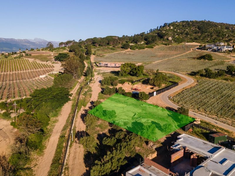 Aerial view of a vacant residential plot highlighted in green, situated in a scenic rural landscape with surrounding vineyards and mountain views.