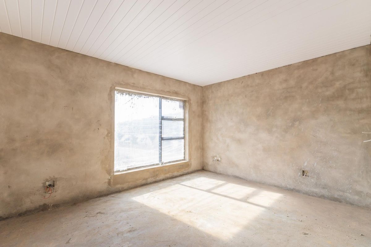 An unfinished bedroom space featuring raw plaster walls, a white tongue and groove ceiling, and a large metal-framed window.