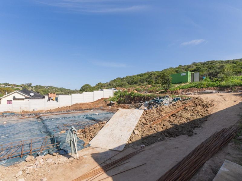 A construction site showing foundation preparation, reinforcing steel, and a precast concrete boundary wall on a sloped plot.