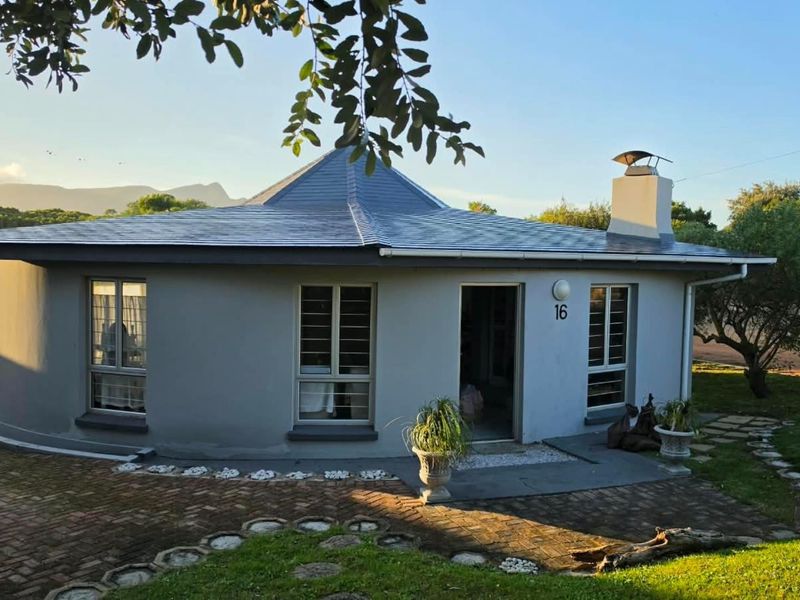 A charming circular-style house featuring a pitched roof, chimney, and paved entrance area, set within a landscaped garden.