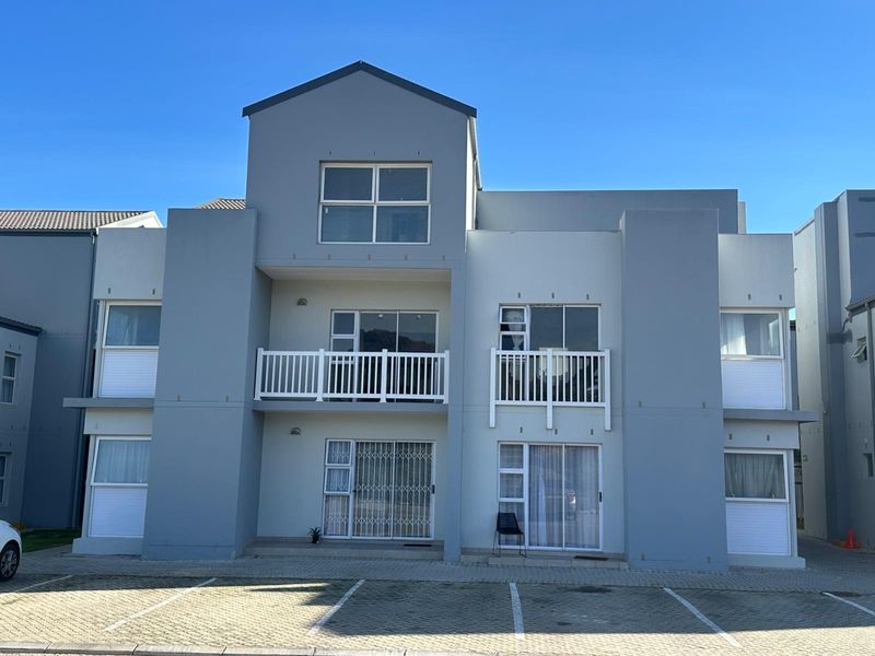 Front elevation of a modern apartment complex featuring paved parking, balconies with white railings, and large sliding glass doors with security gates.