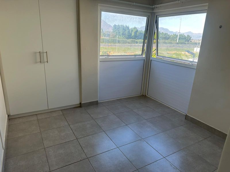 A bright, empty bedroom featuring neutral tiled flooring, built-in cupboards, and large corner windows offering natural light and scenic views.