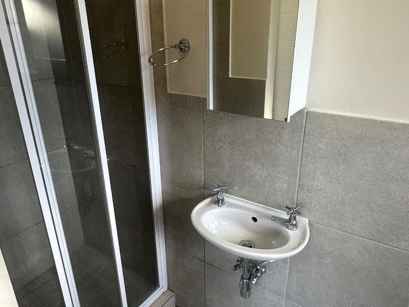 A clean, functional bathroom featuring a glass shower enclosure, wall-mounted basin, and mirrored medicine cabinet with neutral grey wall and floor tiling.
