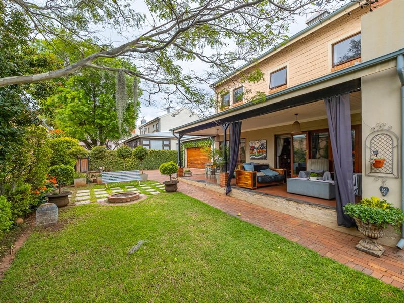 A well-maintained rear garden featuring a covered patio, brick paved walkway, lawn area, and a built-in fire pit.