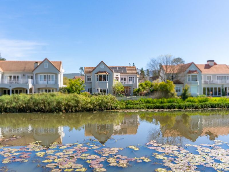 A scenic view of the rear elevation of a residential property featuring a tiled roof, solar panels, and a balcony overlooking a tranquil pond.