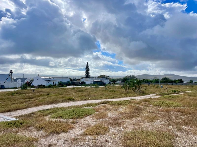 A view of a vacant residential plot featuring natural vegetation and an unpaved access path, set against a cloudy sky.