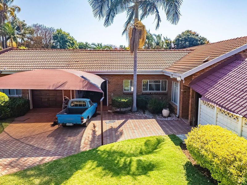 Front elevation of a brick house featuring a paved driveway, covered parking area with a shade sail, and a well-maintained garden.