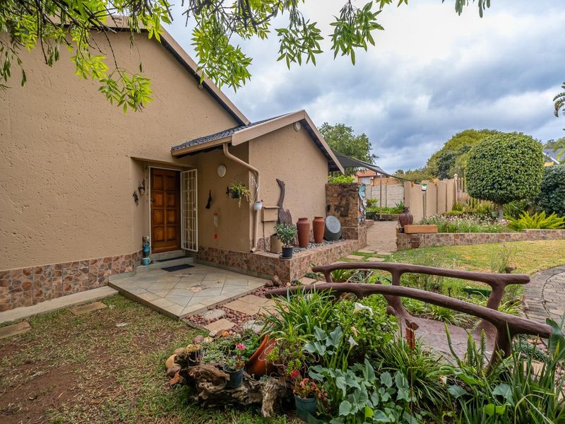 A well-maintained rear exterior view featuring a tiled entrance porch, stone-clad base, and a beautifully landscaped garden with a decorative bridge and paved pathways.