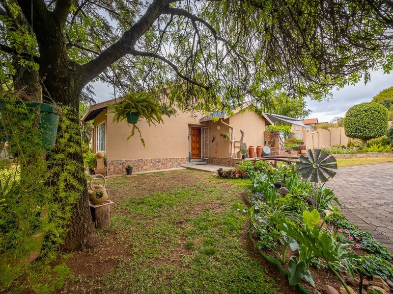 A well-maintained front exterior featuring a paved driveway, stone-clad base, and established garden with mature trees and landscaped flower beds.