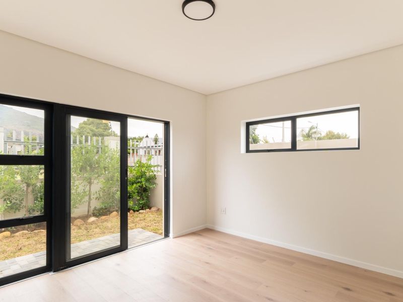 A bright, modern, and empty bedroom featuring light wood laminate flooring, black aluminium window frames, and a sliding door leading to a private garden area.