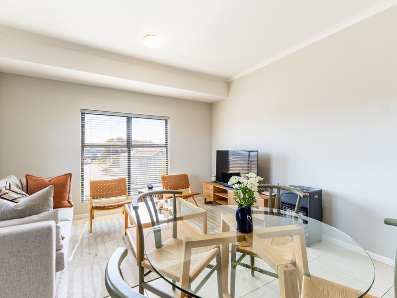 A bright open-plan living and dining area featuring neutral wall tones, tiled flooring, and large windows with venetian blinds.