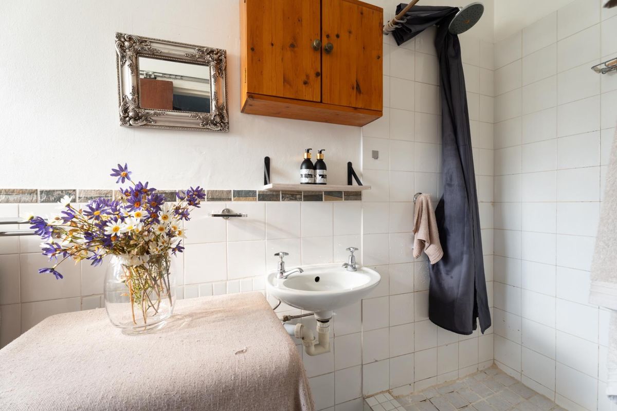 A functional bathroom featuring a wall-mounted basin, tiled shower area, and wooden storage cabinet, requiring some cosmetic updates to modernize the space.
