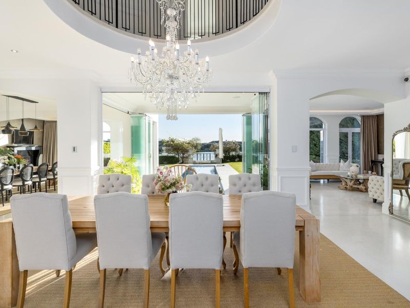 Elegant open-plan dining area featuring a grand crystal chandelier, recessed ceiling detail, and seamless flow through stacking glass doors to the pool area.