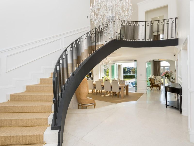 Grand entrance hall featuring a sweeping curved staircase with wrought iron balustrade, elegant wainscoting, and a statement crystal chandelier, leading into the open-plan living area.