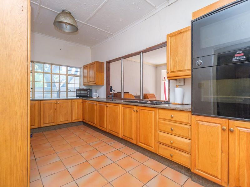 A dated kitchen featuring wooden cabinetry, built-in oven, hob, and tiled flooring, with a convenient pass-through window connecting to the adjacent living space.