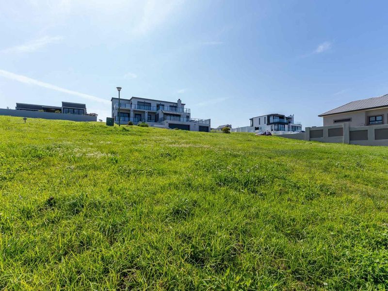 A view of a sloped, undeveloped residential plot with surrounding boundary walls and neighboring modern homes in the background.