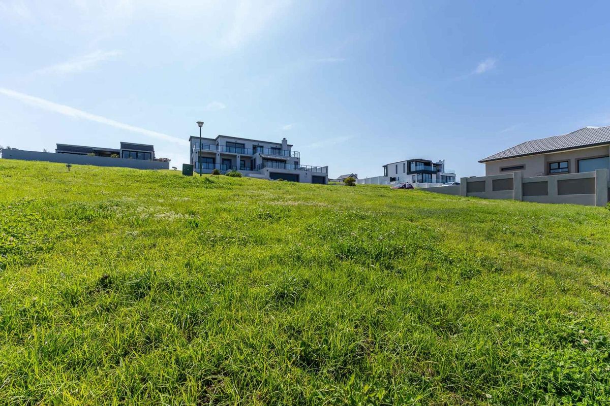 A view of a sloped, undeveloped residential plot with surrounding boundary walls and neighboring modern homes in the background.
