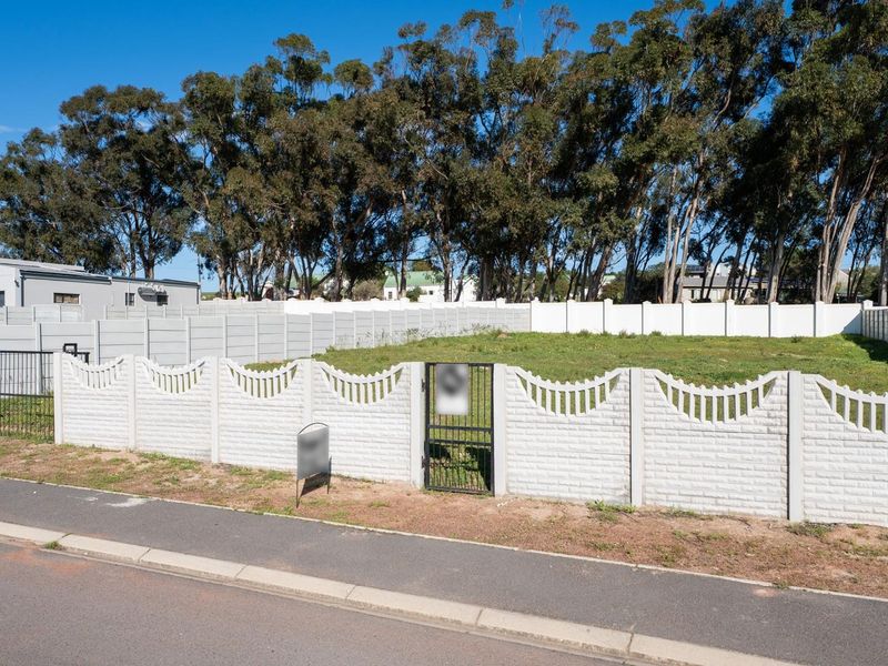 A vacant residential plot enclosed by a white precast concrete boundary wall with a metal pedestrian gate, situated along a paved street.