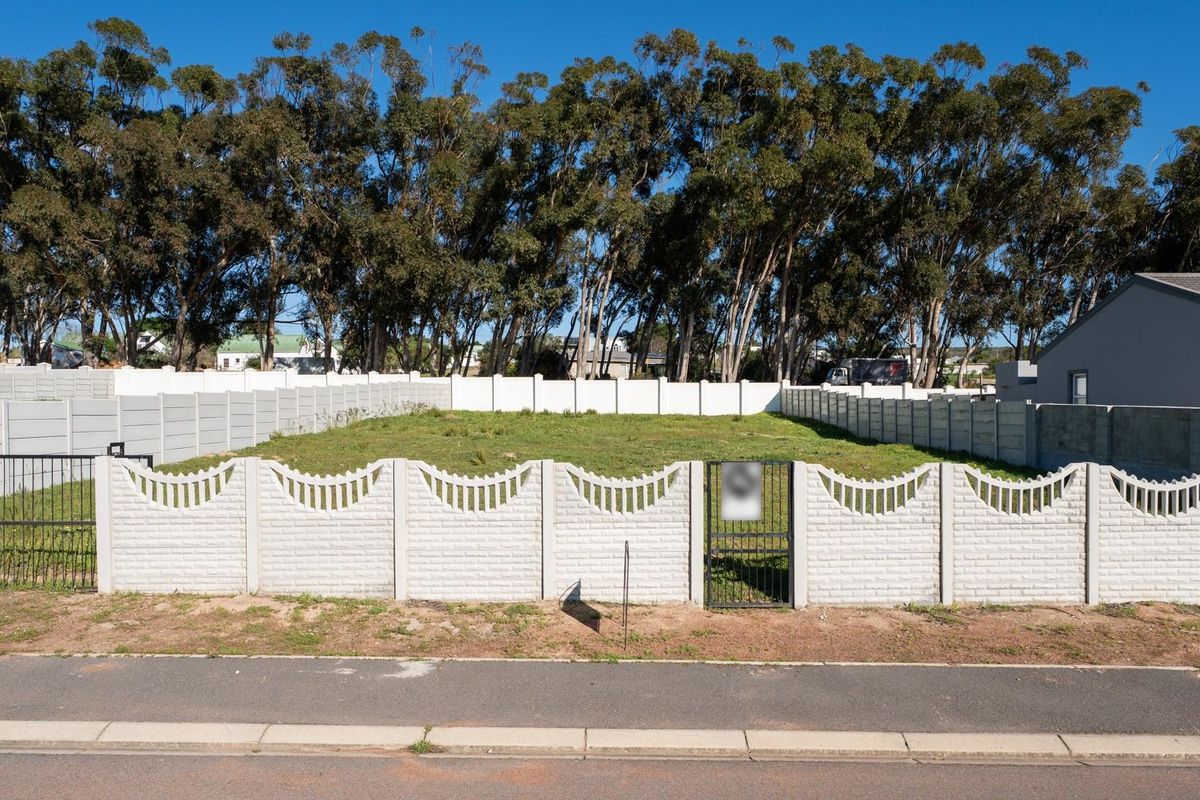 A vacant residential plot enclosed by a precast concrete boundary wall with a metal pedestrian gate, featuring established trees along the rear boundary.