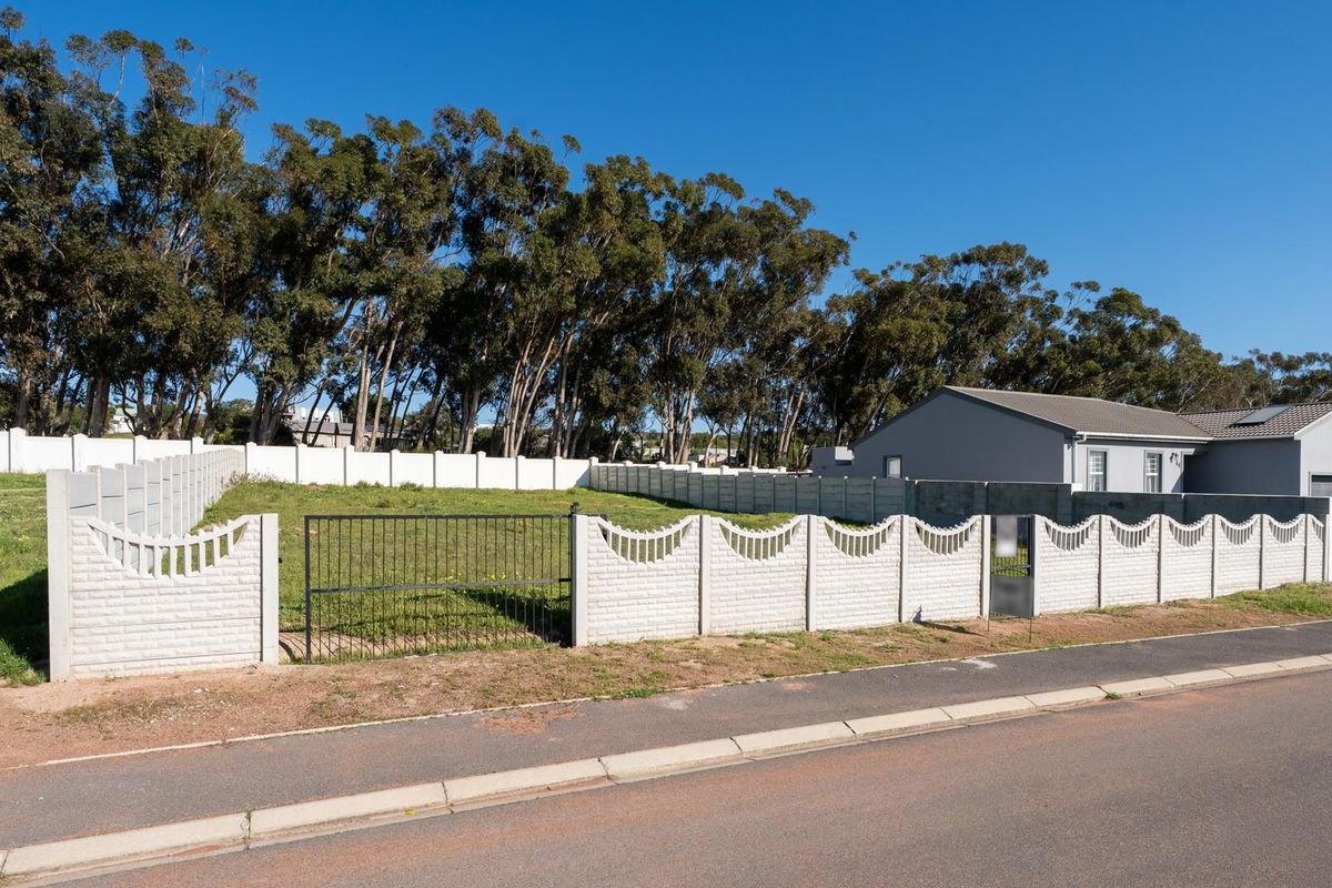 A vacant residential plot enclosed by a precast concrete boundary wall and metal gate, situated along a paved street with established trees in the background.