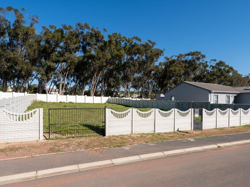 A vacant residential plot enclosed by a precast concrete boundary wall and metal gate, situated along a paved street with established trees in the background.